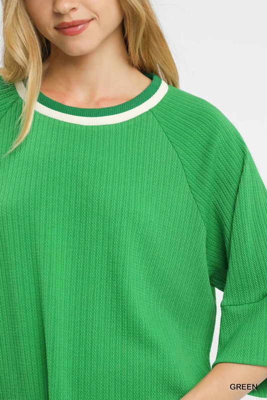 A close-up of a woman wearing a bright green ribbed knit top with a white contrast trim around the crew neckline. The image is cropped to show the upper torso, neck, and lower face of the model against a plain light background.