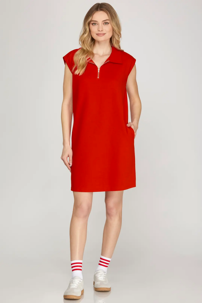 A woman modeling a bright red sleeveless polo-knit dress with a short zipper at the collar, side pockets, and a relaxed fit, paired with white sneakers and red-and-white striped socks, against a light gray studio background.