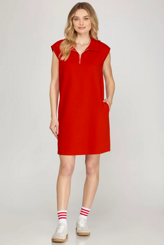 A woman modeling a bright red sleeveless polo-knit dress with a short zipper at the collar, side pockets, and a relaxed fit, paired with white sneakers and red-and-white striped socks, against a light gray studio background.
