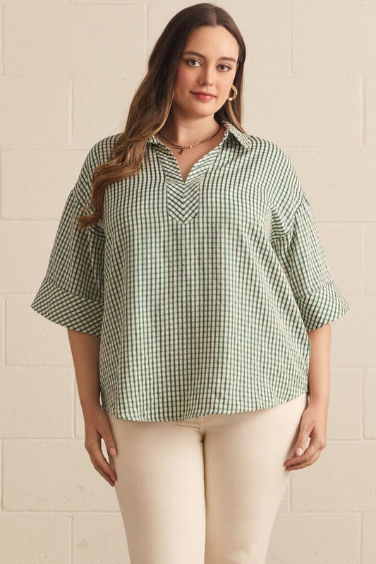 A woman modeling a green and white gingham check short-sleeve button-down shirt with a split V-neck, paired with cream pants, standing against a light beige brick wall.