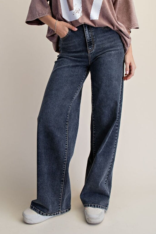 A person wearing dark blue wide-leg denim jeans with a faded wash, styled with a dusty rose graphic t-shirt and white sneakers against a light beige studio background.