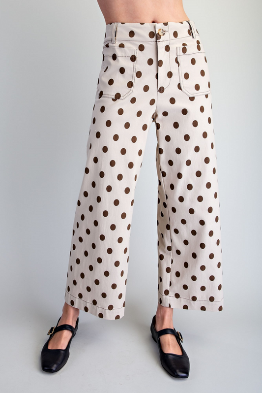 A person wearing high-waisted, wide-leg pants with a brown polka dot pattern on a cream background, paired with black Mary Jane shoes, shown from the waist down against a light gray studio background.