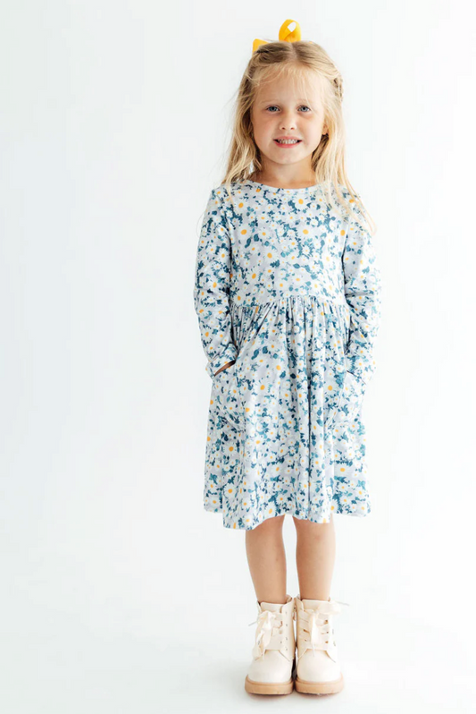 A young girl with blonde hair and a small yellow bow stands against a plain white background wearing a long-sleeved knee-length dress with a blue, white, and yellow floral pattern, and cream-colored lace-up boots.
