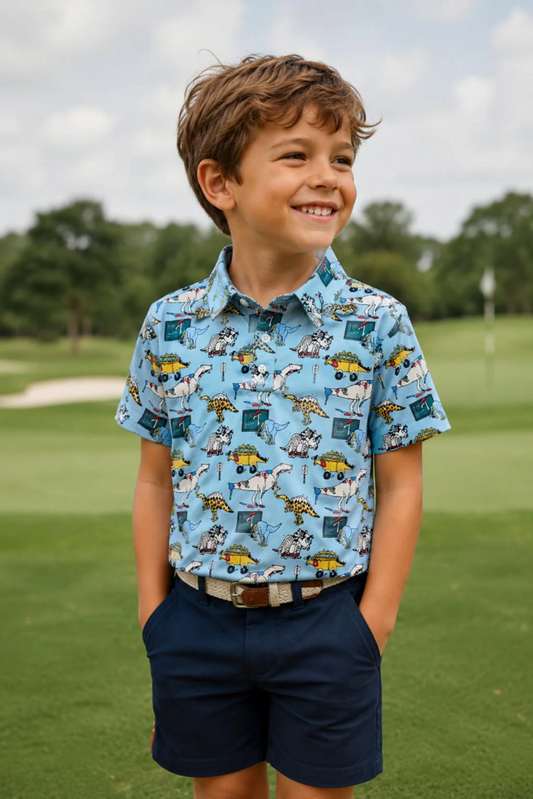 A young boy smiling on a golf course, wearing a light blue short-sleeve button-up shirt with a cartoon construction worker pattern and navy blue shorts.