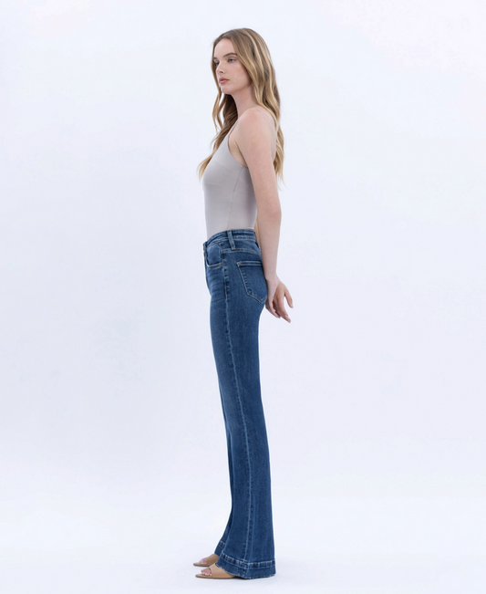 A woman stands in profile wearing blue high-waisted flare jeans and a light beige sleeveless top against a clean white studio background.