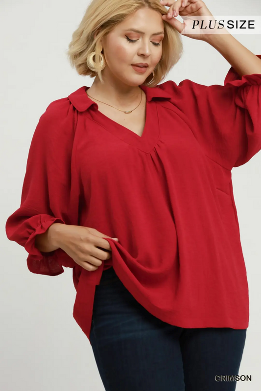 A woman wearing a red plus-size blouse with a V-neck, long sleeves, and a gathered front, paired with dark blue jeans. The brand name 'PLUS SIZE' is visible in the top right corner.