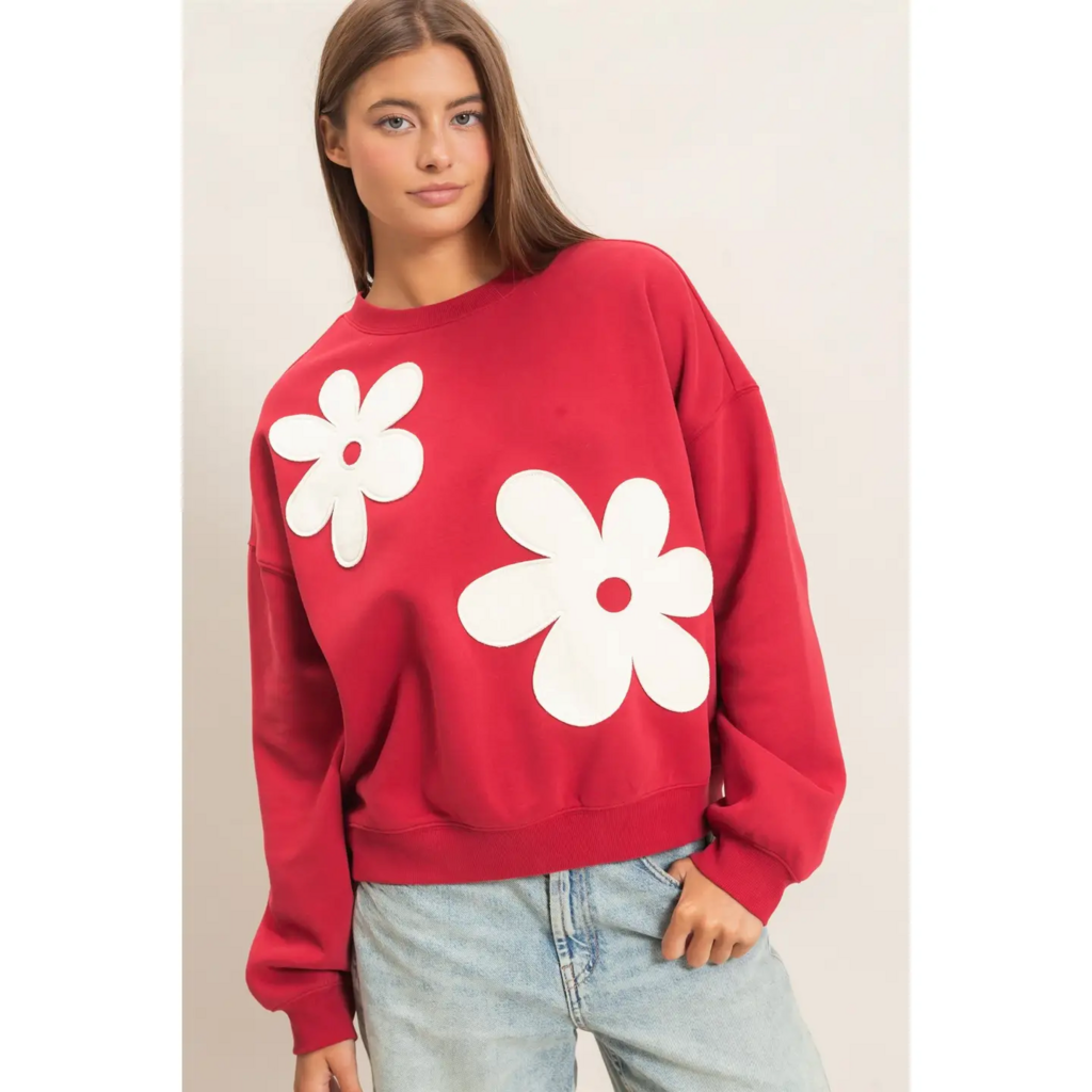 A woman wearing a red crewneck sweatshirt with two large white flower appliqués on the front, paired with light-wash jeans, against a plain light beige studio background.