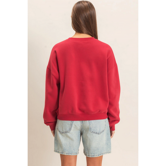 A woman viewed from the back wearing a bright red crewneck sweatshirt and light-wash denim shorts against a plain beige studio background.