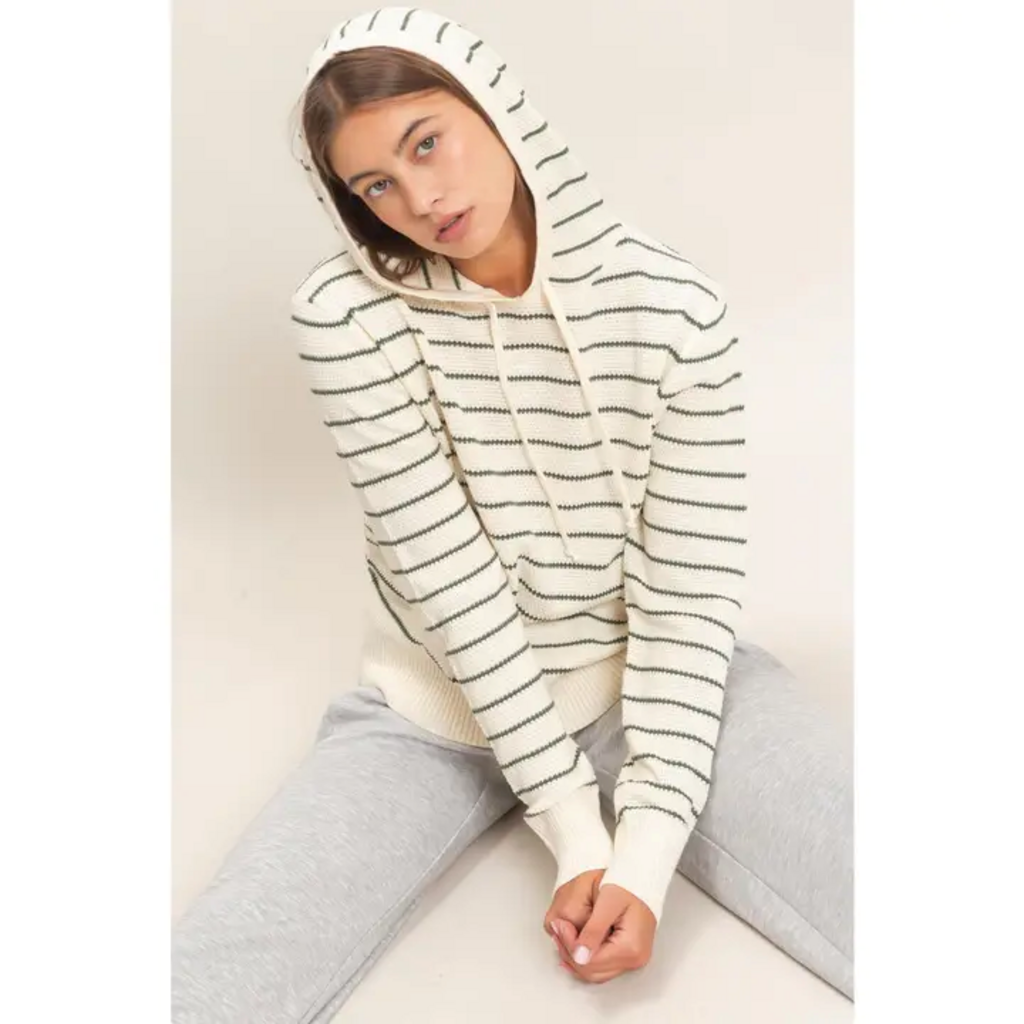 A woman wearing a cream-colored knit hoodie with thin dark green horizontal stripes, paired with light gray sweatpants, sitting against a plain beige background.